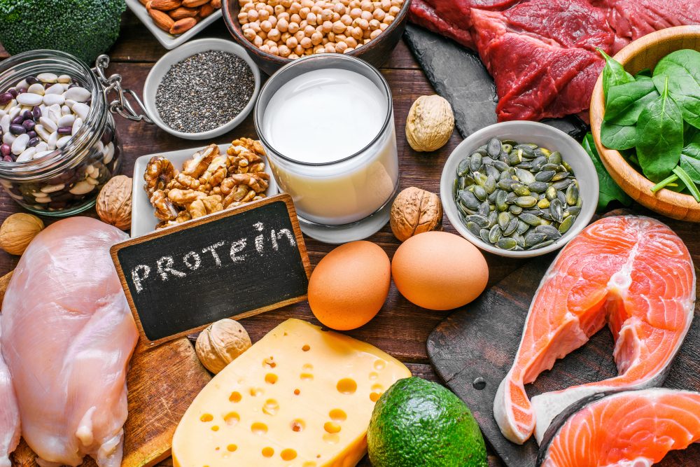 A variety of high-protein foods including chicken, beef, salmon, eggs, cheese, nuts, seeds, dairy, beans, and leafy greens, with a small chalkboard labeled 