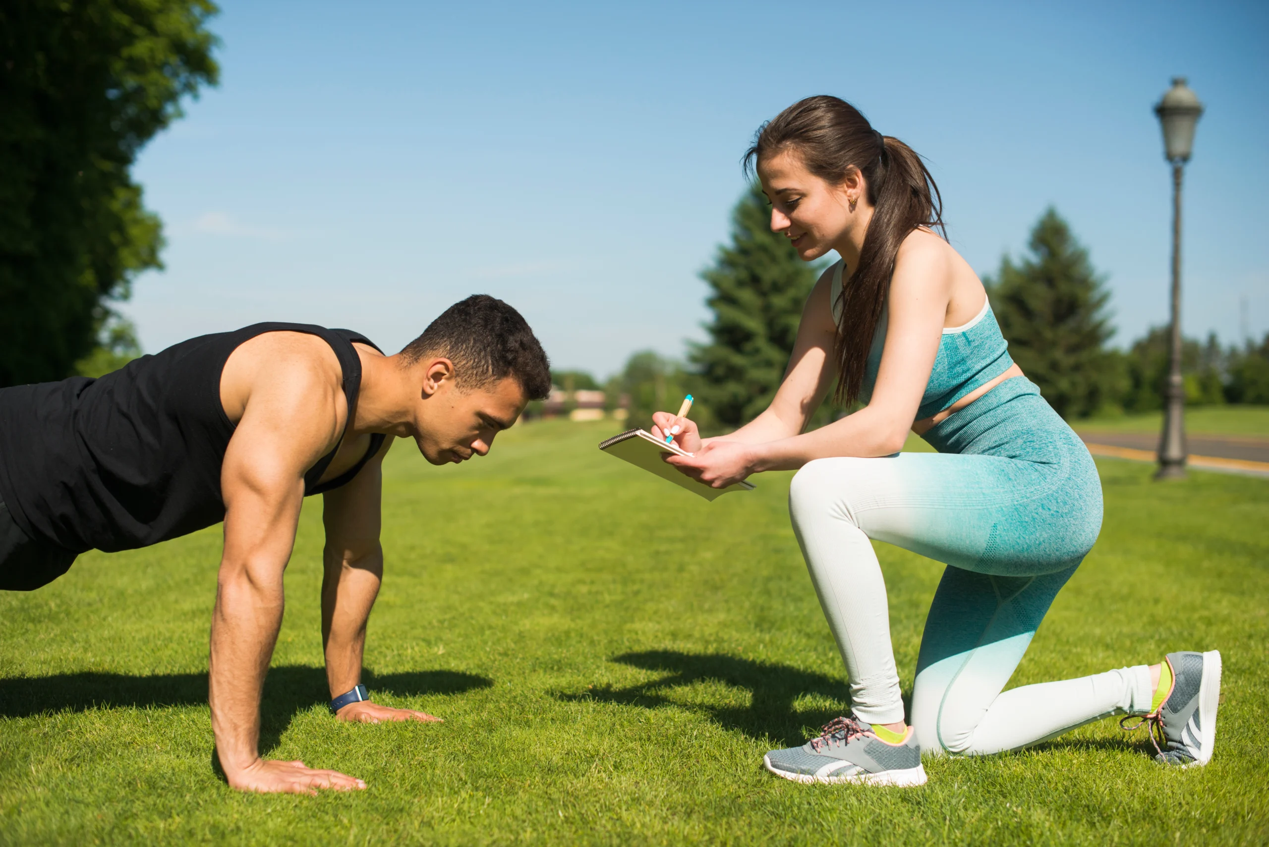 A man doing a plank exercise on the grass while a woman in athletic wear kneels beside him, taking notes on a clipboard.